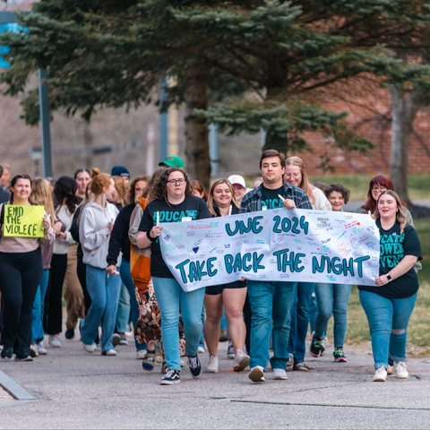 A group of students marches on campus holding a sign that reads "ֱ 2024 Take Back the Night"